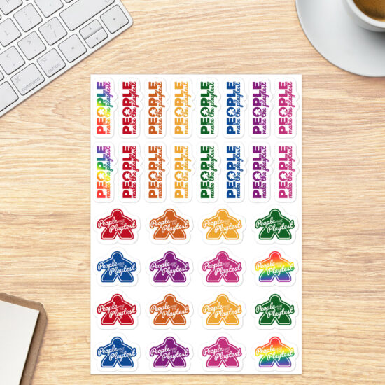 Sheet of multicolor People Make the Playtest Stickers, 16 with an outer shape of a meeple and 16 with a meeple in the O of "People" shown on a tabletop next to a coffee cup and keyboard for scale