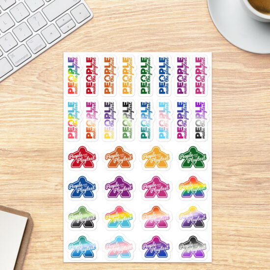 Sheet of LGBTQ pride and rainbow colored People Make the Playtest Stickers, 16 with an outer shape of a meeple and 16 with a meeple in the O of "People" shown on a tabletop next to a coffee cup and keyboard for scale